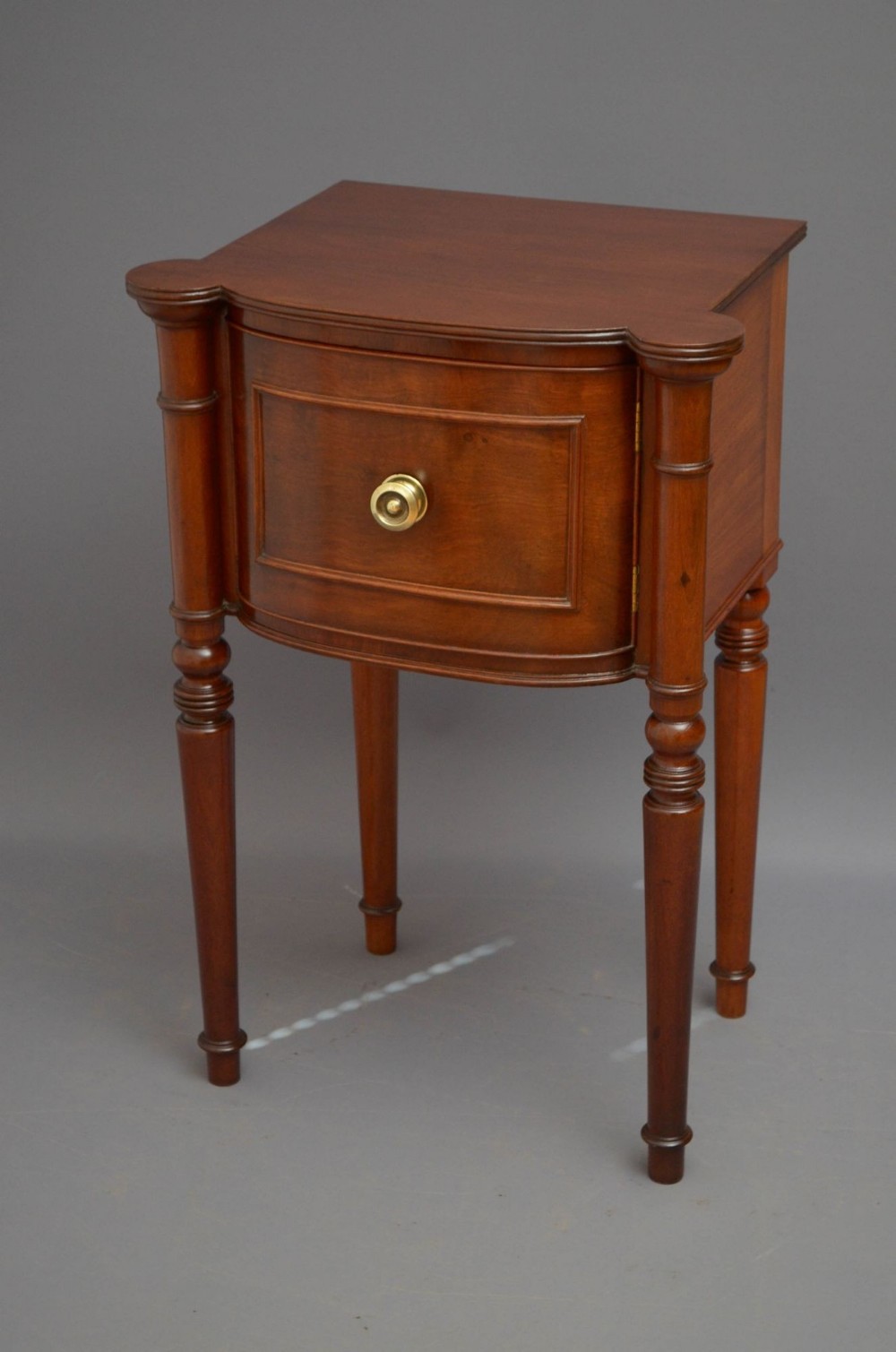 A Regency period mahogany bedside cabinet featuring turned column supports, a curved front panel with a brass knob, and tapered legs.
