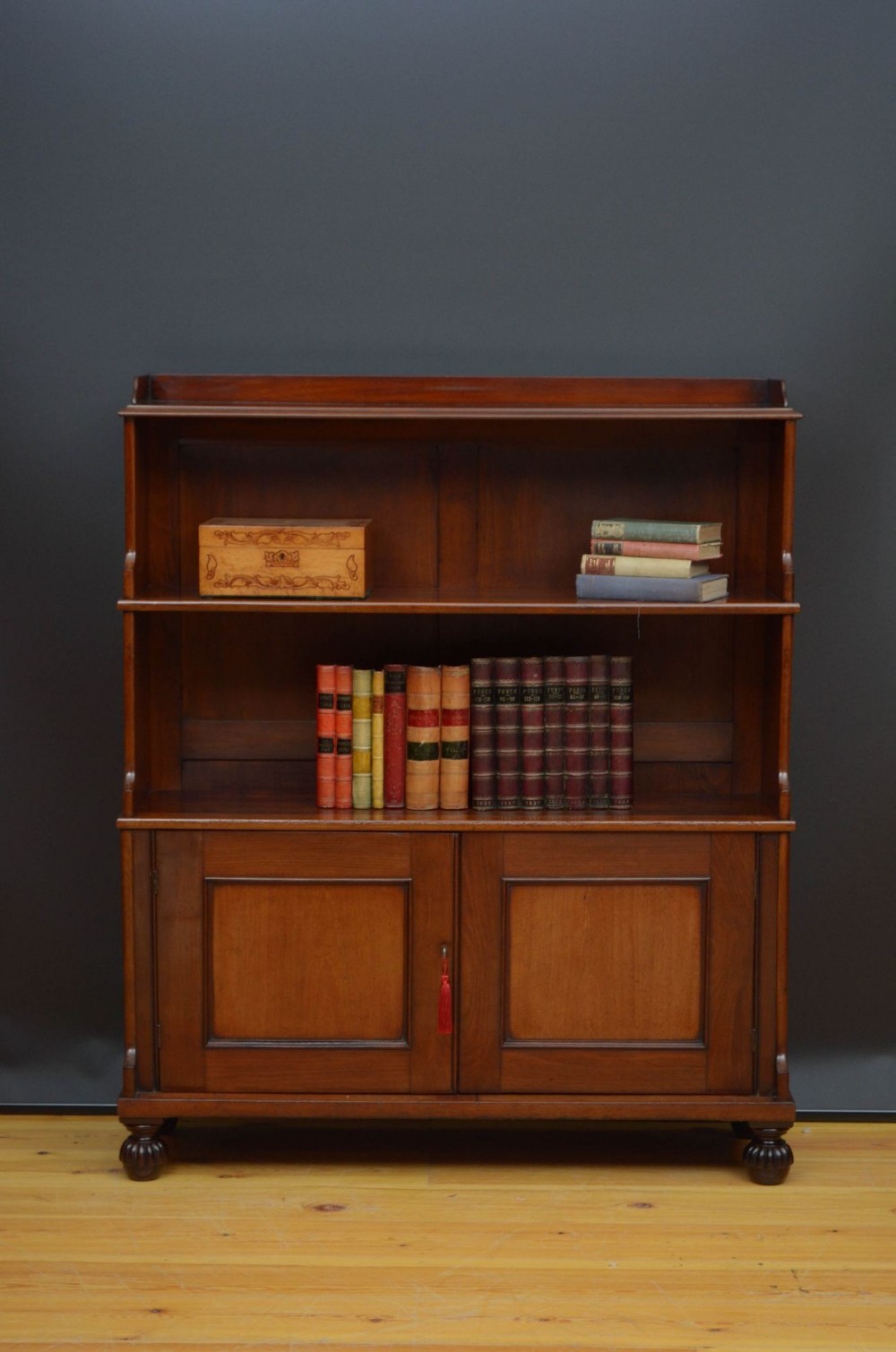 A William IV mahogany waterfall bookcase featuring two open graduated upper shelves over a double-door lower cabinet resting on turned bun feet.