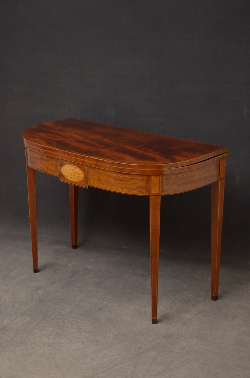 A Regency D-shaped mahogany tea table featuring a fold-over top, central satinwood oval inlay, and square tapered legs.