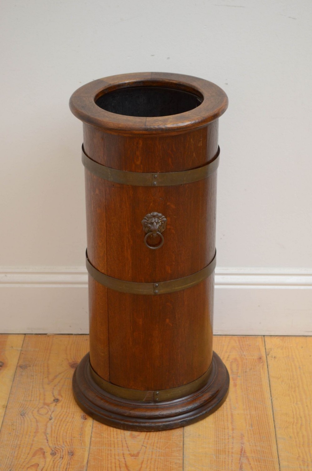 A cylindrical 19th-century oak coopered stick stand featuring brass binding hoops, a central lion mask drop handle, and a moulded circular base.