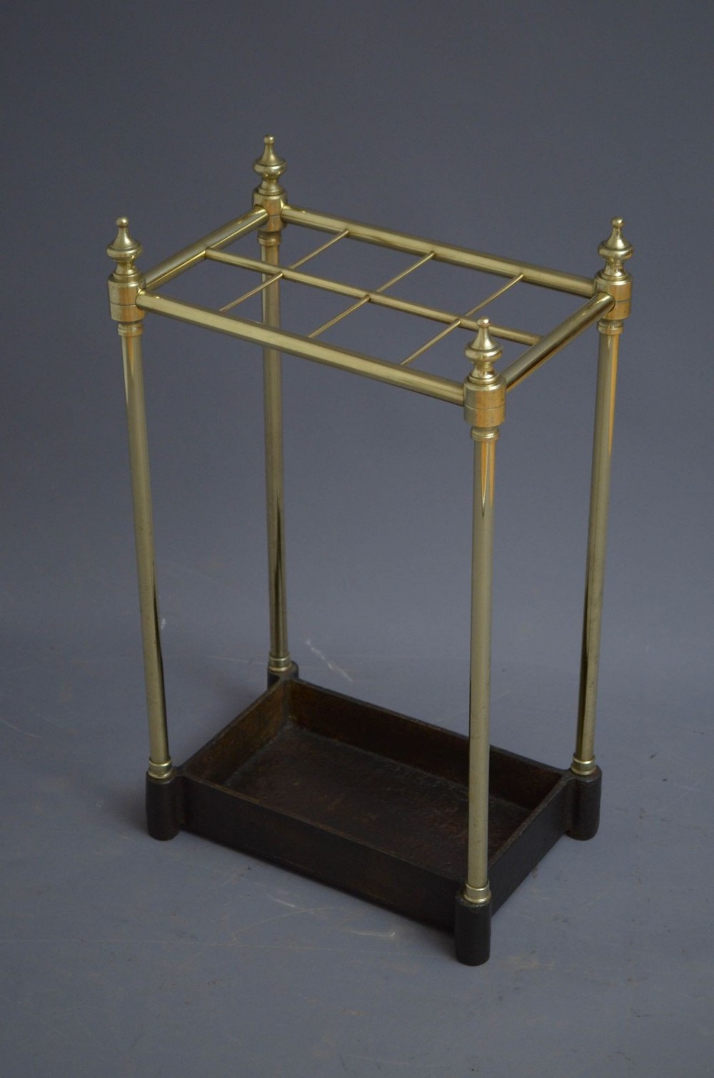 A Victorian brass umbrella stand featuring a rectangular tubular frame with four urn finials, six divided sections, and a dark cast iron drip pan base.