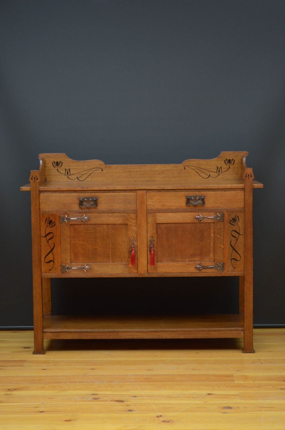 An Arts and Crafts solid oak sideboard featuring stylised floral inlays, copper strap hinges, two drawers above a two-door cabinet, and an open lower shelf.
