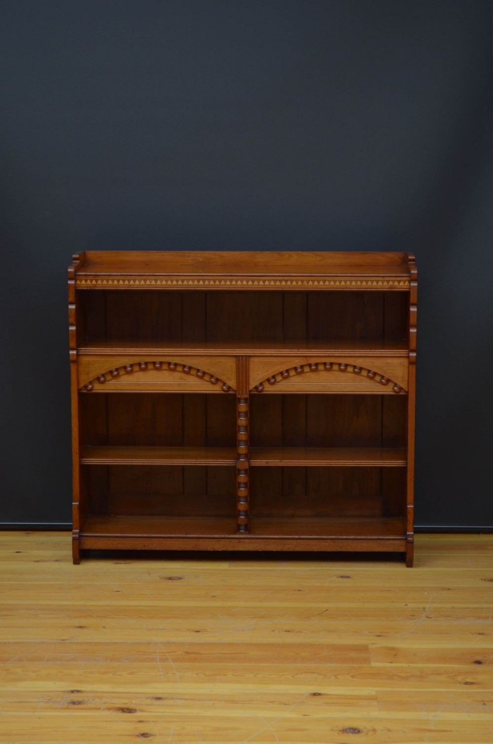 An Aesthetic Movement warm brown walnut open bookcase featuring carved arched detailing, geometric frieze inlay, and central turned spindles.