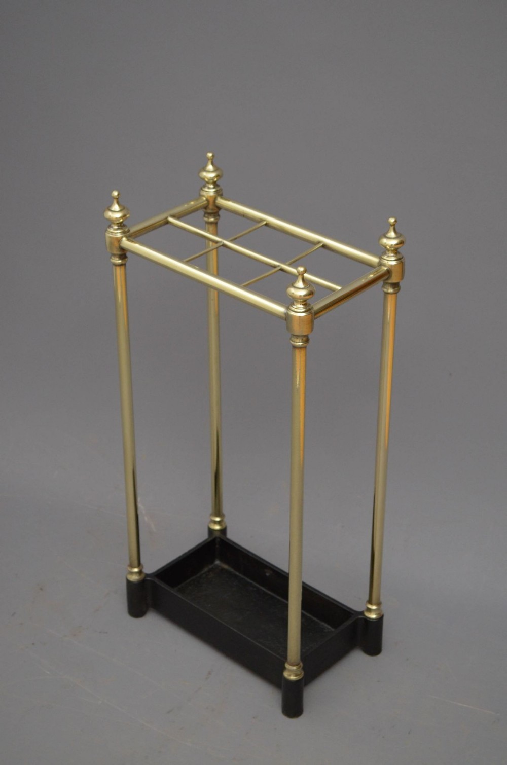 A Victorian brass umbrella stand featuring four vertical columns topped with finials, six divided top compartments, and a black cast iron drip tray at the base.