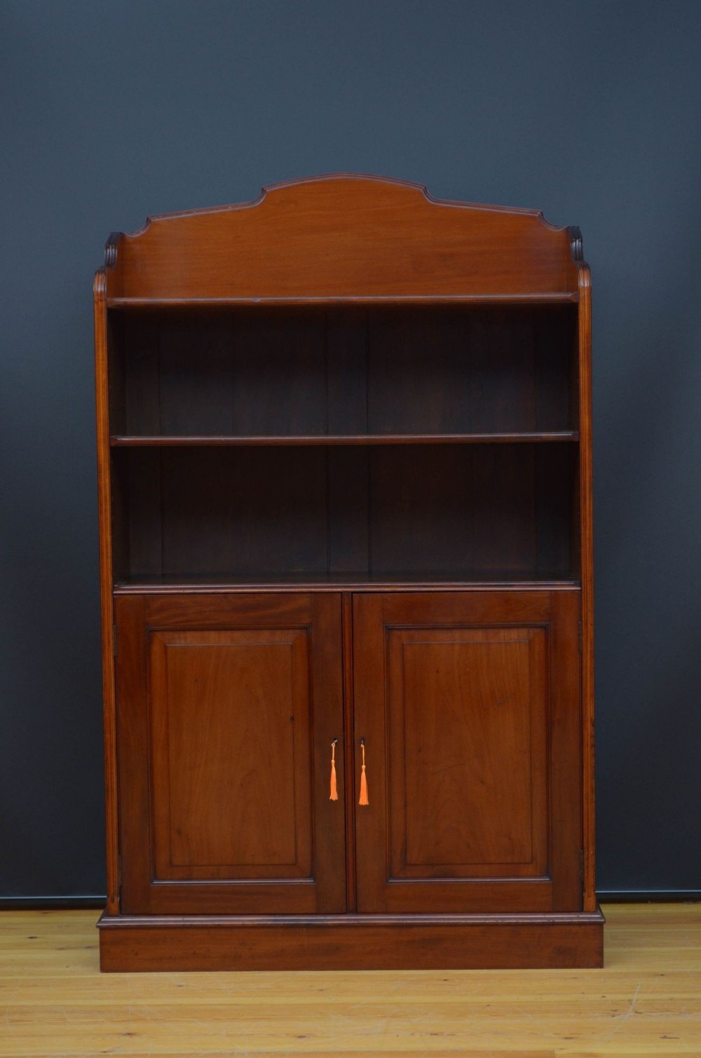 A solid mahogany Victorian open bookcase featuring a shaped cresting rail, two open upper shelves, and a double-door lower cupboard with panelled fronts.