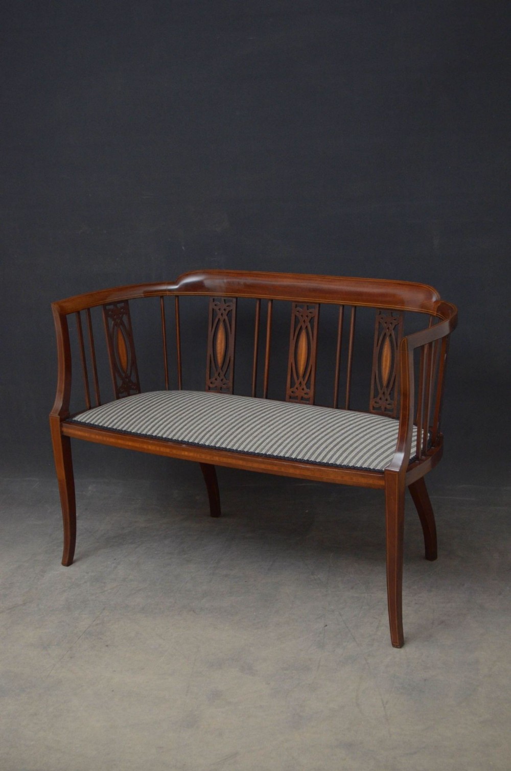 An Edwardian mahogany settee featuring a curved backrest with inlaid wood splats, slim vertical spindles, and a striped upholstered seat supported by tapered legs.