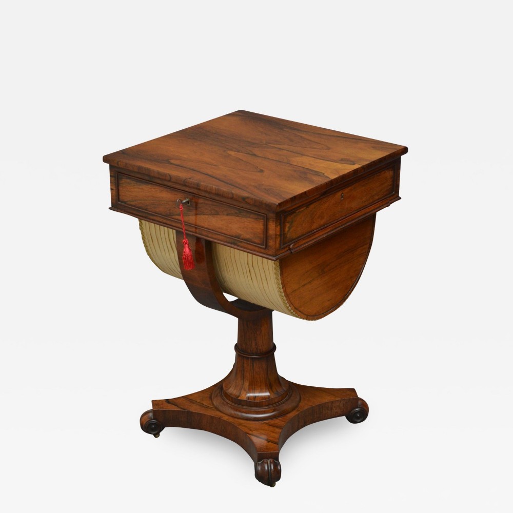 A 19th-century William IV rosewood work and writing table featuring a square top with a frieze drawer, a pleated fabric-covered work basket, and a turned pedestal on a concaved base with scroll feet.