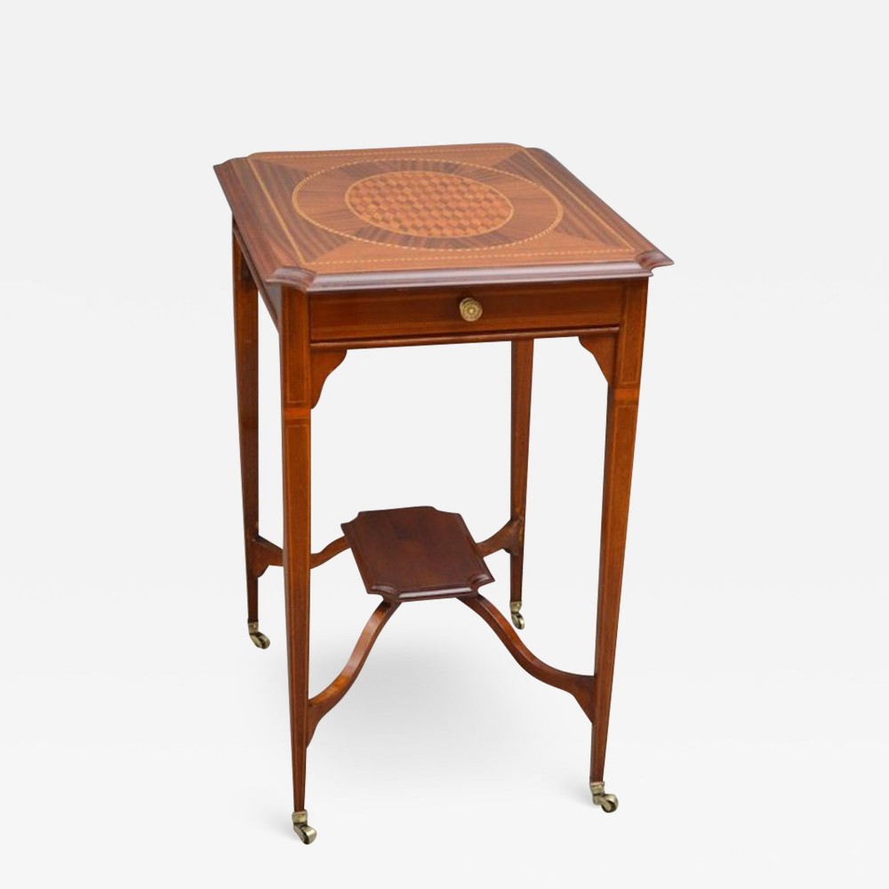 An Edwardian inlaid mahogany occasional table featuring a square top with marquetry geometric patterns, a single drawer, slender tapered legs with line stringing, a lower undertier, and brass castors.