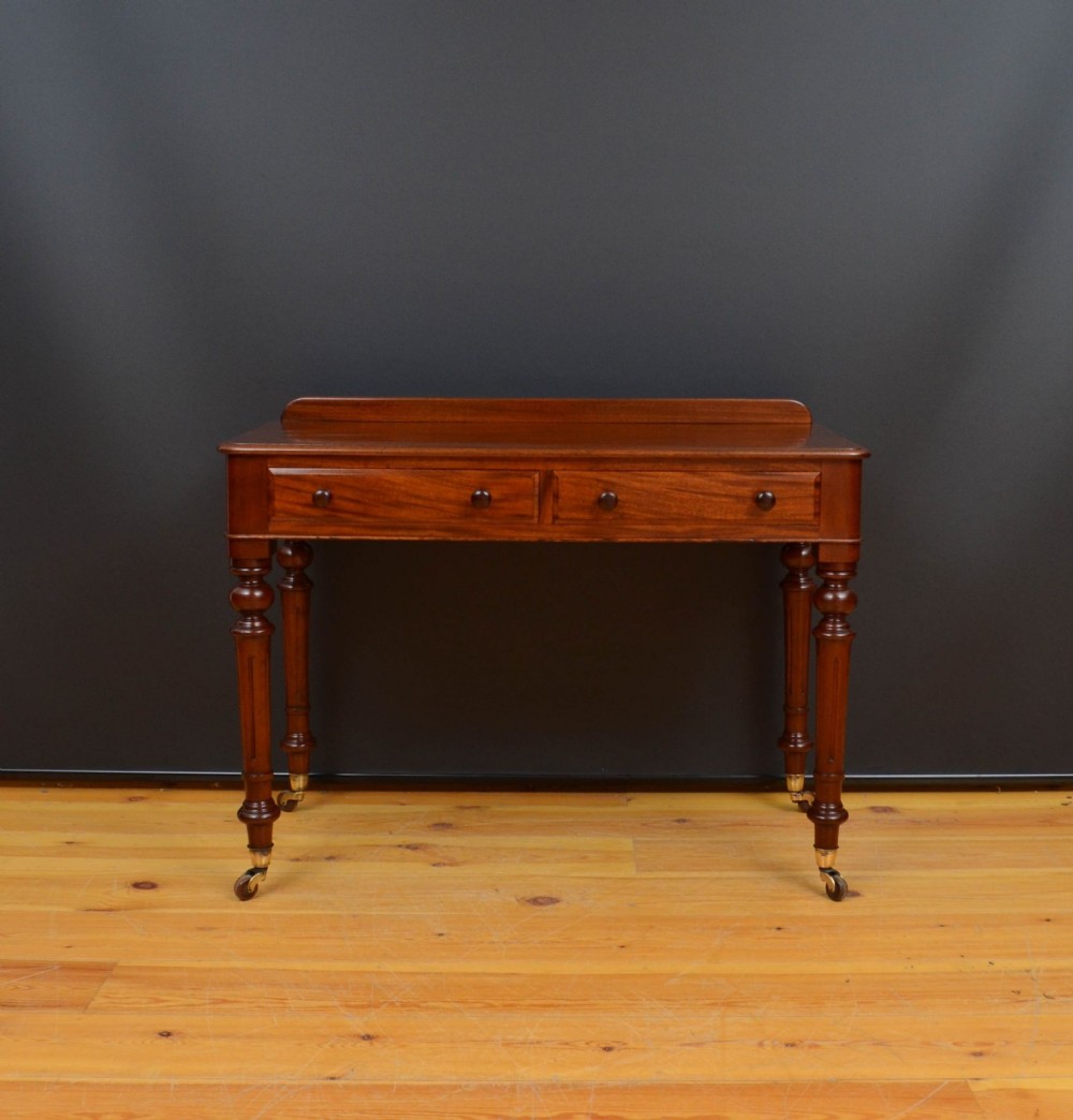 A Victorian solid mahogany writing table featuring a low rear upstand, two frieze drawers with round wooden knobs, and four turned legs on brass castors.