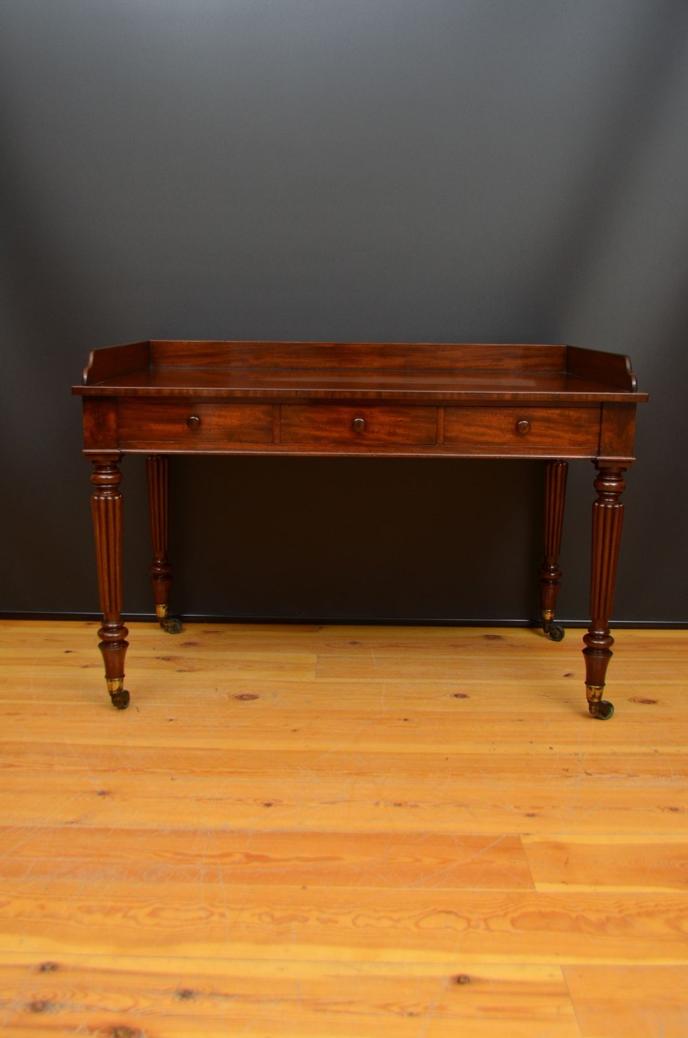 A mahogany writing table featuring three frieze drawers, a raised rear gallery upstand, reeded turned legs, and brass castors.