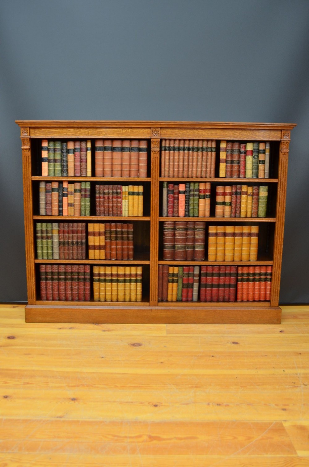 A wide Victorian oak open bookcase featuring two bays with four tiers of shelves filled with books and carved floral pilaster details.
