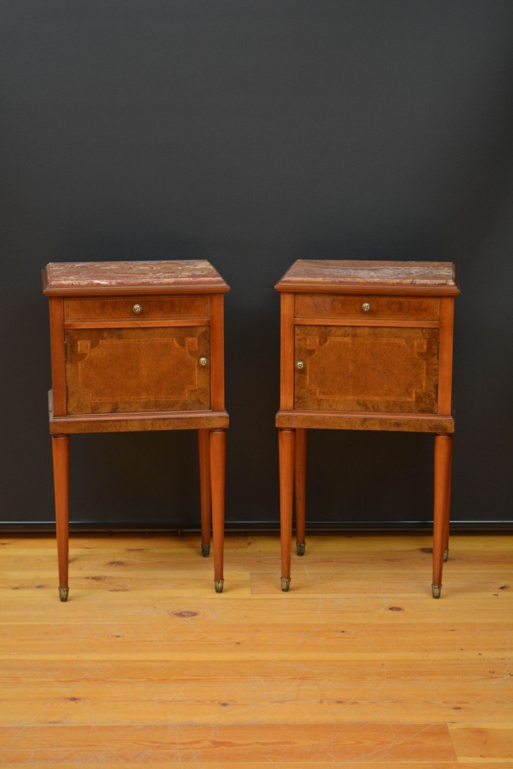 A pair of tall, square wooden bedside cabinets with marble tops, geometric inlaid panels, upper drawers, lower cupboards, and slim tapered legs with brass feet.