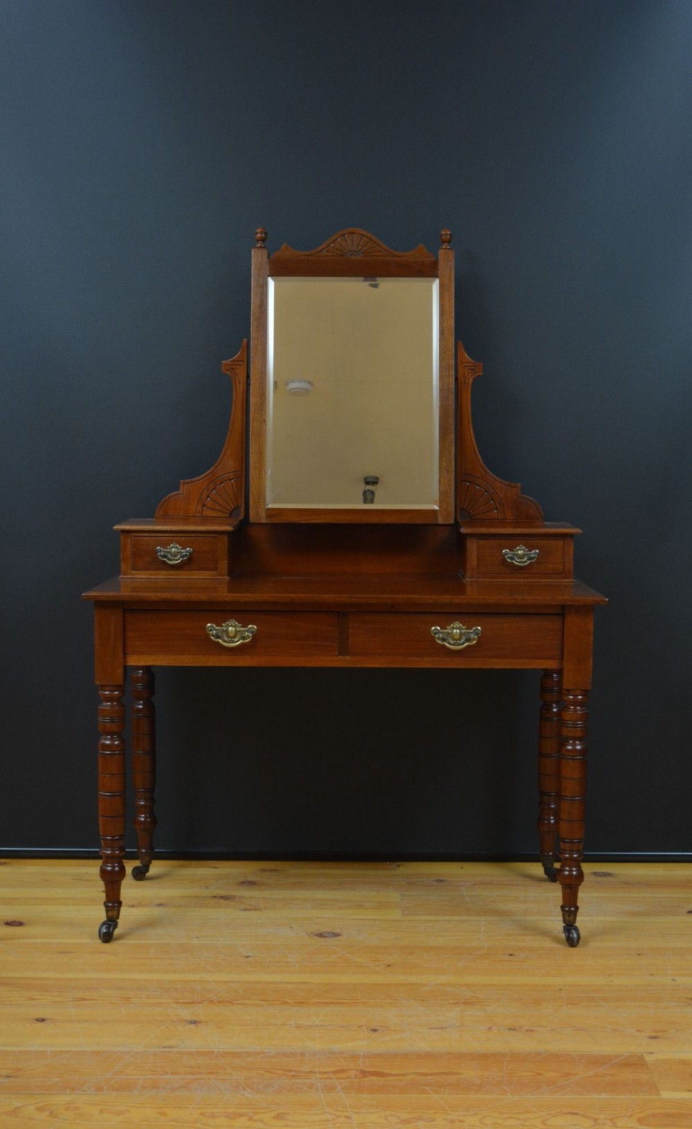 An Edwardian mahogany dressing table featuring a rectangular central swivel mirror, fan-carved pediments, two small trinket drawers, two lower desk drawers with brass handles, and turned legs on castors.