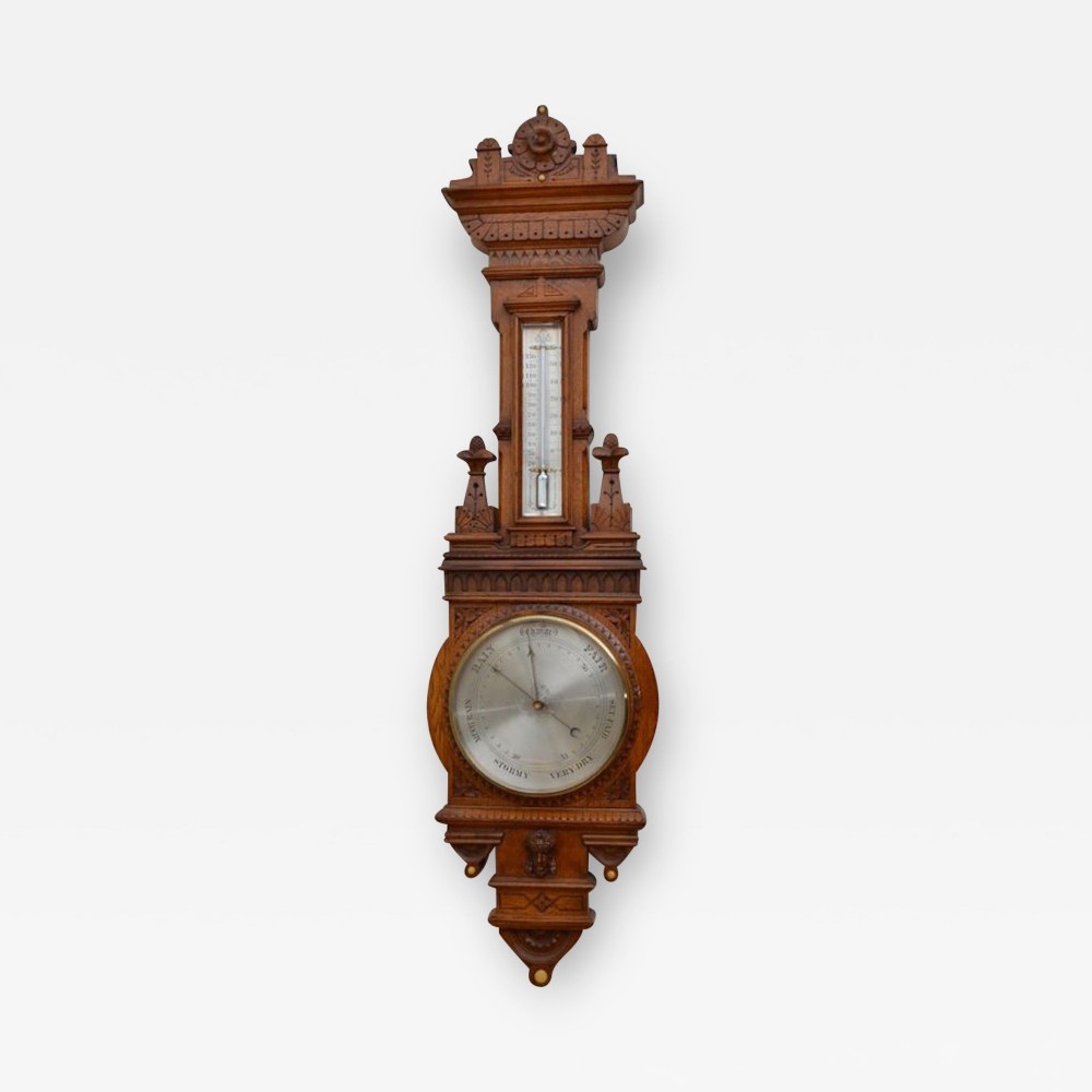 A Victorian carved oak aneroid wall barometer featuring a vertical thermometer tube positioned above a circular silvered dial with black lettering.