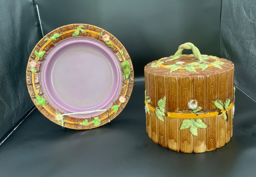 A Victorian George Jones majolica cheese keep featuring a wooden stave texture, moulded green leaves, pink flowers, and a twisted vine handle, alongside a matching base plate with a lilac interior glaze.