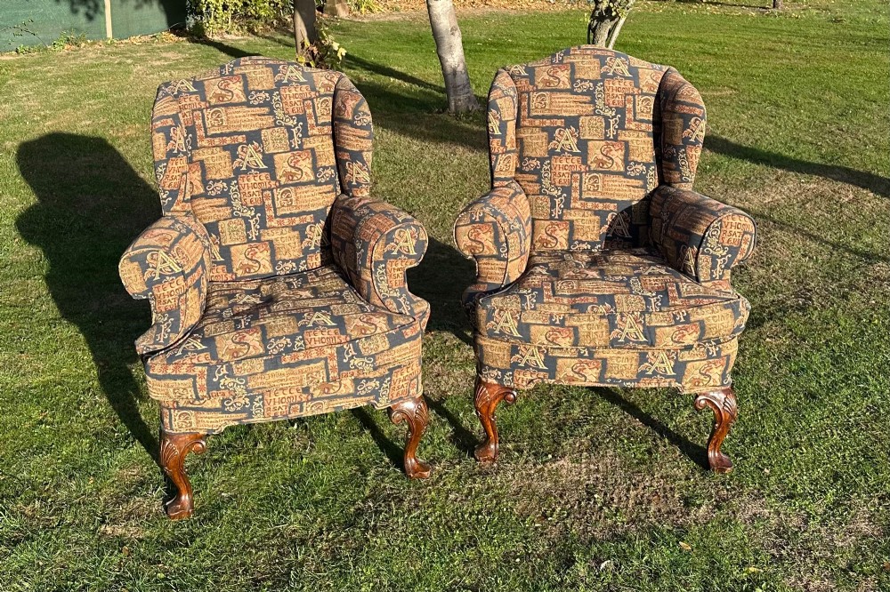 A pair of 1920s wingback armchairs upholstered in a gold and blue manuscript patterned tapestry fabric with carved wooden cabriole front legs.