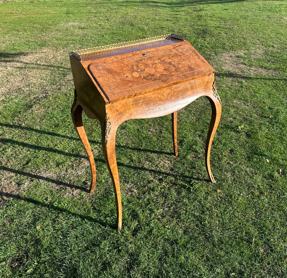 A 19th-century French bureau de dame crafted with floral marquetry inlay, slender cabriole legs, decorative brass corner mounts, and a brass gallery along the top.