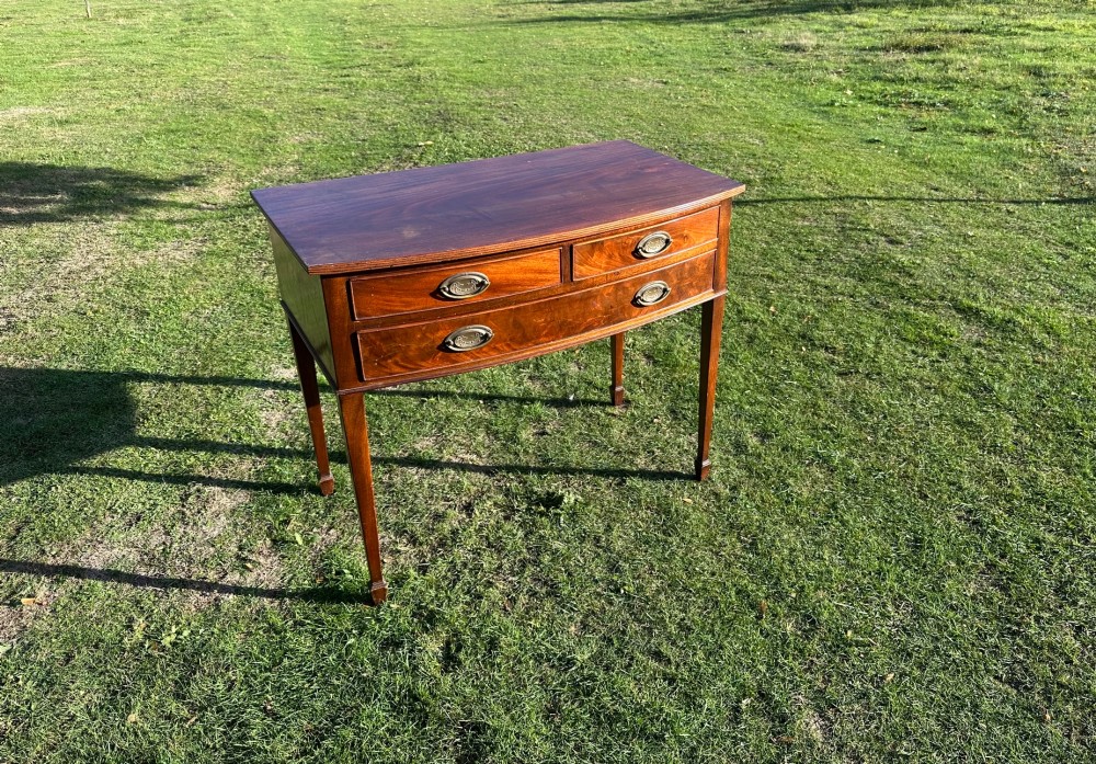 A circa 1920 mahogany bow front side table featuring three drawers with brass oval drop handles and slender tapered legs.