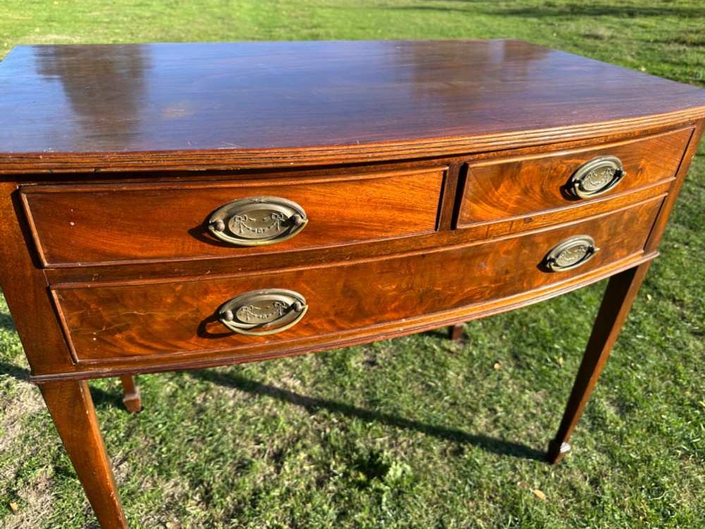 A circa 1920 mahogany bow front side table featuring three drawers with brass oval drop handles and slender tapered legs. View II