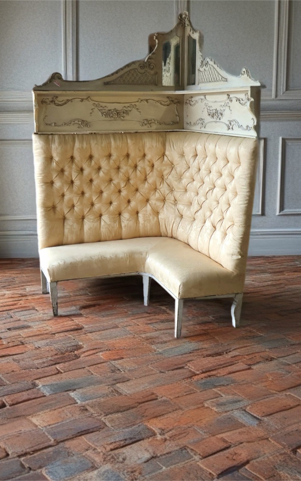 A high-backed 1920s French corner sofa with cream button-tufted upholstery, carved cream-painted timber framing, and a top crest featuring inset mirror panels.