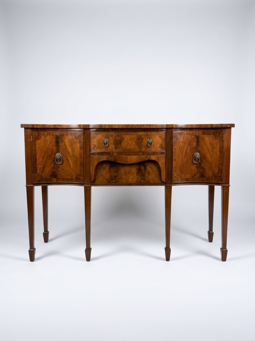 A Georgian serpentine-front mahogany sideboard with flame mahogany veneered doors, square tapered legs, and brass lion-mask drop ring handles.