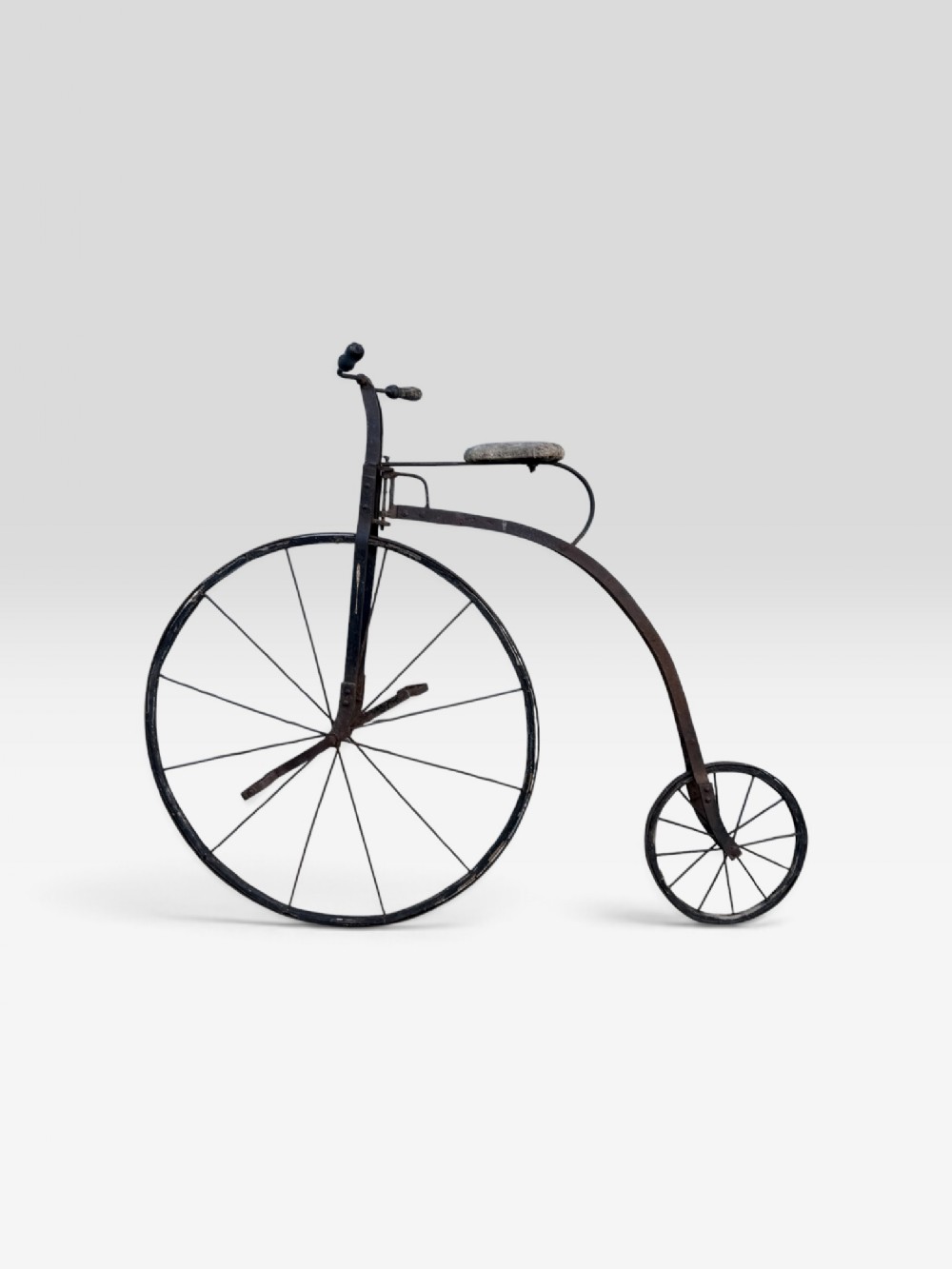 A 19th-century childs penny farthing bicycle featuring a large spoked front wheel, small rear wheel, dark wrought iron frame, and padded seat.
