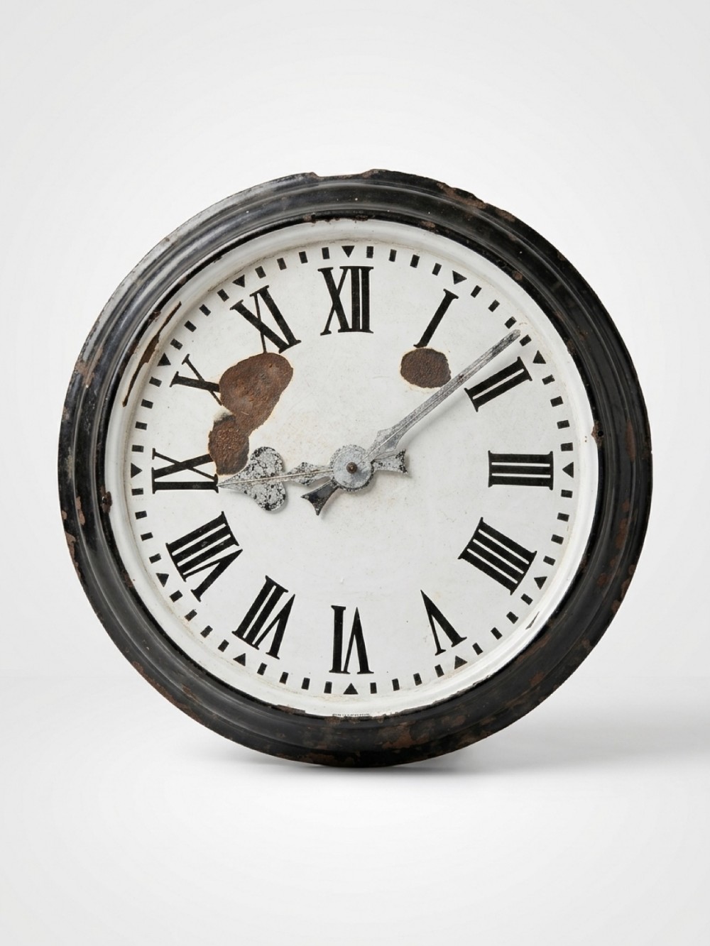 A circular vintage wall clock featuring a white enamelled dial with black Roman numerals, spade hands, and a patinated dark metal bezel with chipped enamel detailing.