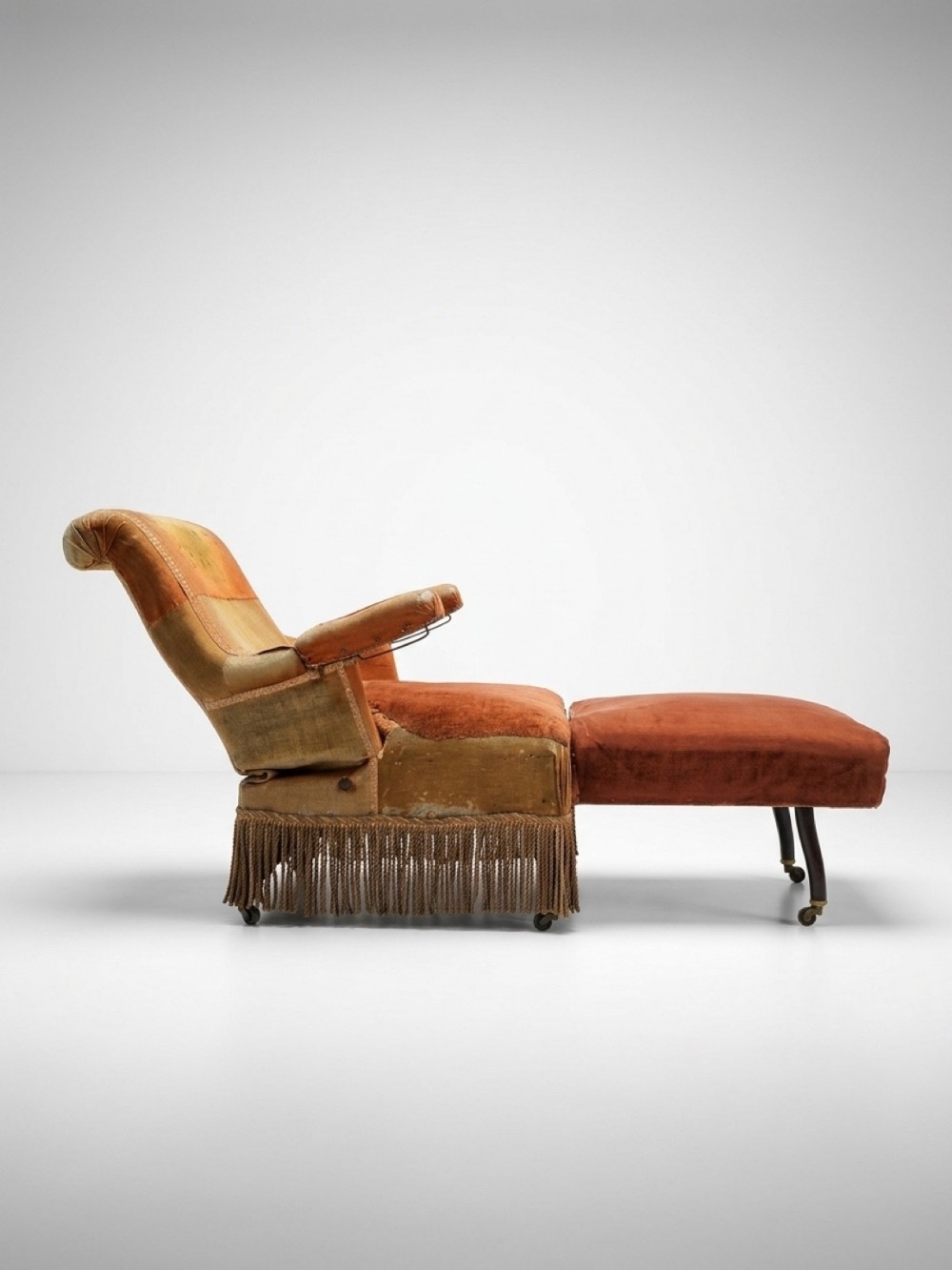 A nineteenth-century French reclining daybed upholstered in worn terracotta velvet with fringe trim and brass castors.