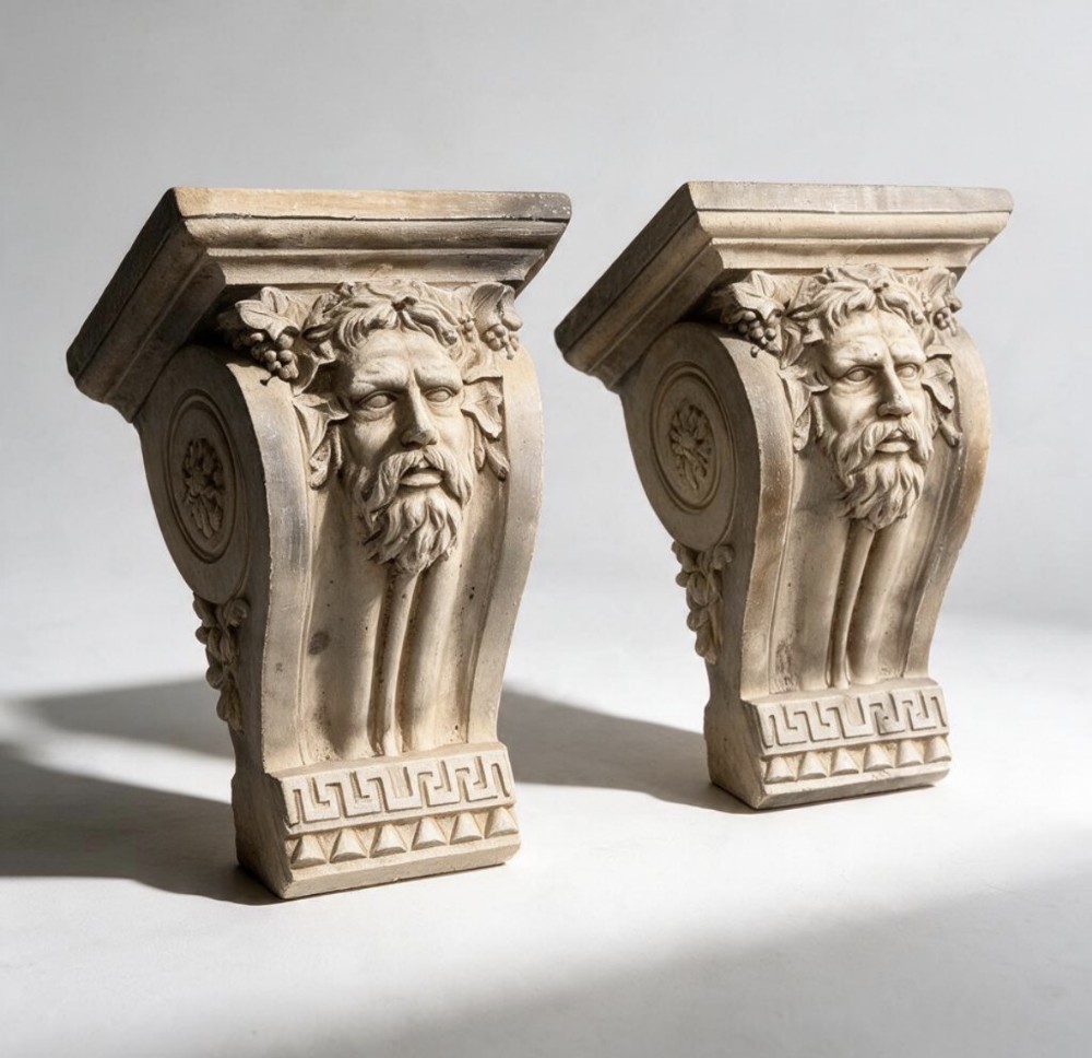 A pair of off-white plaster corbels featuring carved bearded masks surrounded by grape leaves and Greek key base details.