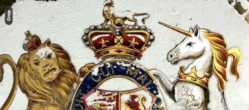 An Edwardian heraldic shield-shaped plaque featuring the gilded British Royal Coat of Arms with a lion, unicorn, and mottoes against a painted off-white background with distressed gold trim. View II