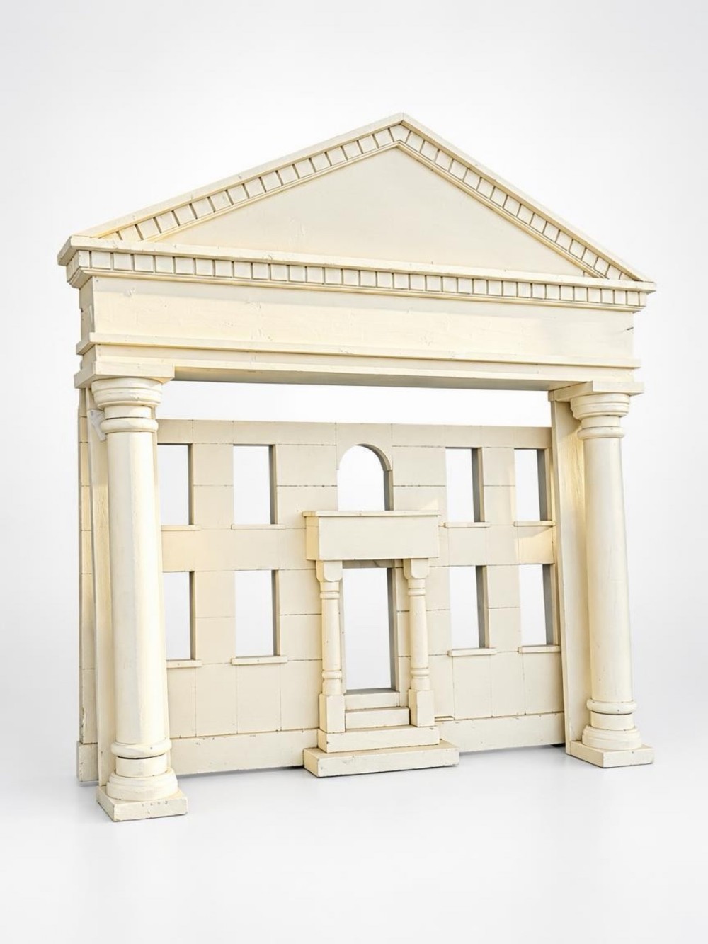 A cream-painted architectural model facade featuring classical columns, a triangular pediment, rectangular window openings, and a central doorway.