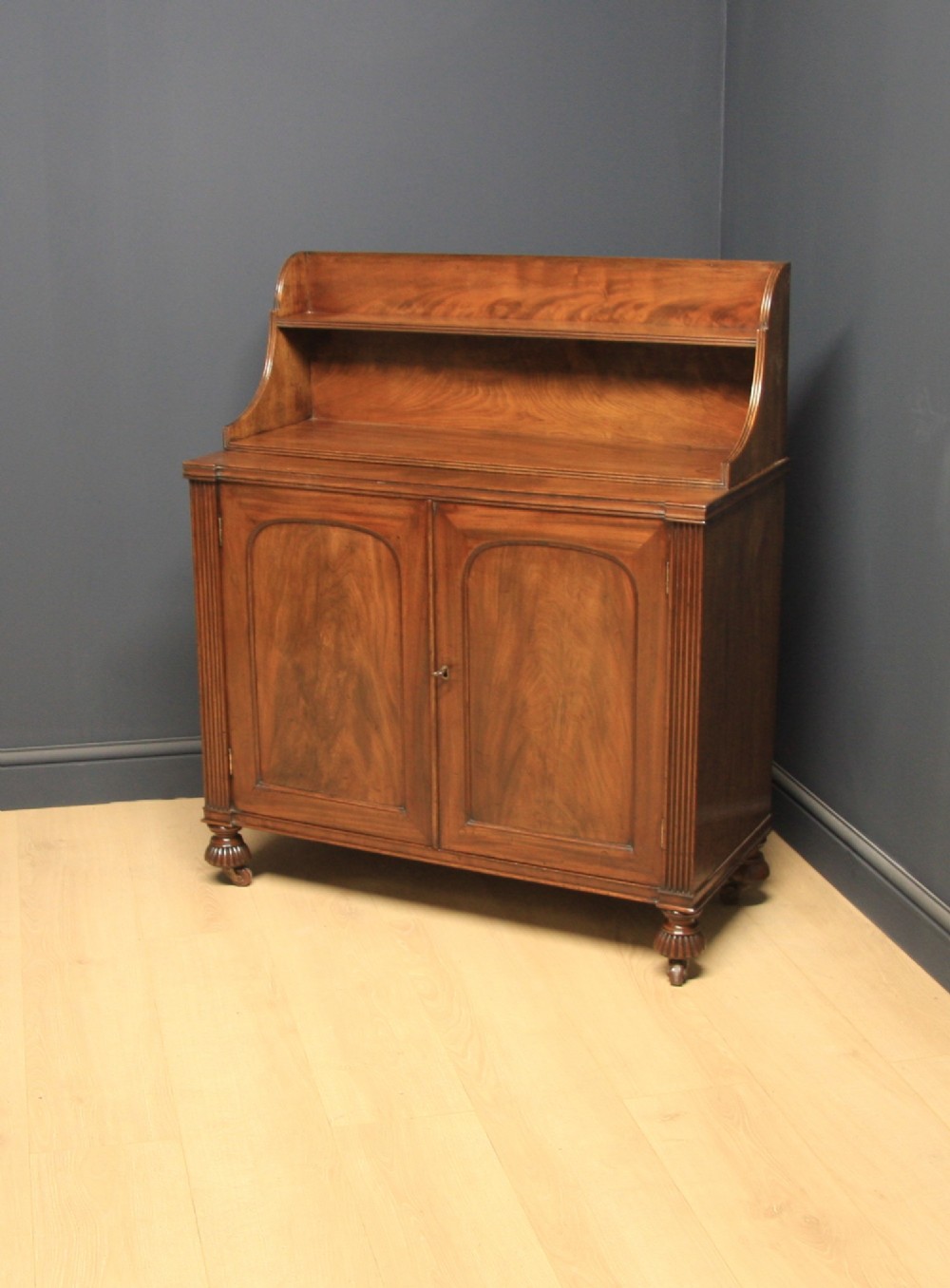 A William IV mahogany sideboard featuring a raised back shelf, flame mahogany panelled cupboard doors, reeded side pilasters, and carved turned feet.