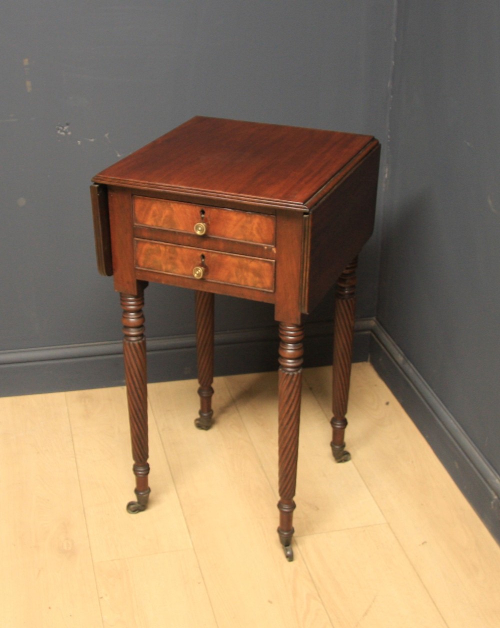 A Regency mahogany drop-leaf table featuring two drawers with lighter wood fronts, round brass knobs, and spiral-turned legs terminating in brass castors.