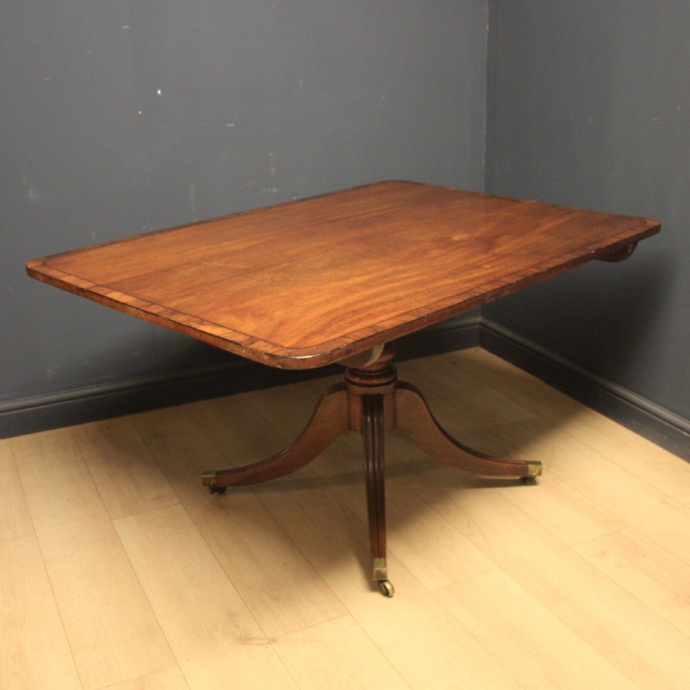 A Georgian mahogany pedestal dining table featuring a rectangular crossbanded top supported by a central turned column and four splayed legs with brass castors.