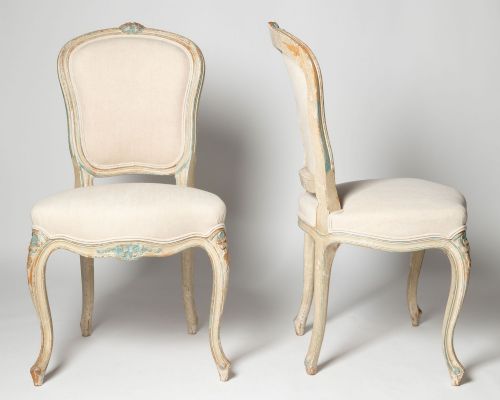 fig 1. A pair of antique French side chairs featuring distressed off-white painted frames with floral crest carvings, cabriole legs, and neutral linen upholstery.