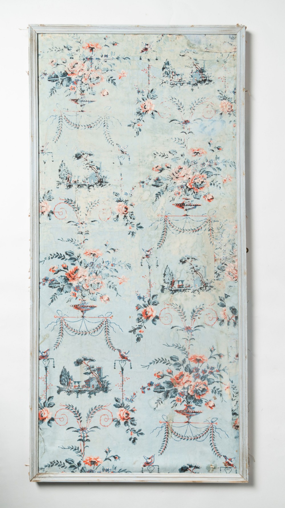 A tall rectangular panel containing 18th-century block-printed pale blue wallpaper with floral urns, swags, and pastoral scenes inside a grey wooden frame.