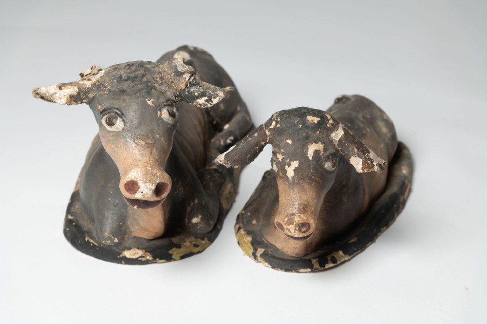 A pair of distressed antique papier-mache cow figurines shown in a resting recumbent position with black, brown, and cream hand-painted finish.