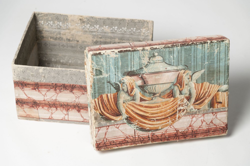 An open rectangular antique French paper box featuring a lithographed lid depicting classical winged figures and an urn, with patterned paper lining the interior.