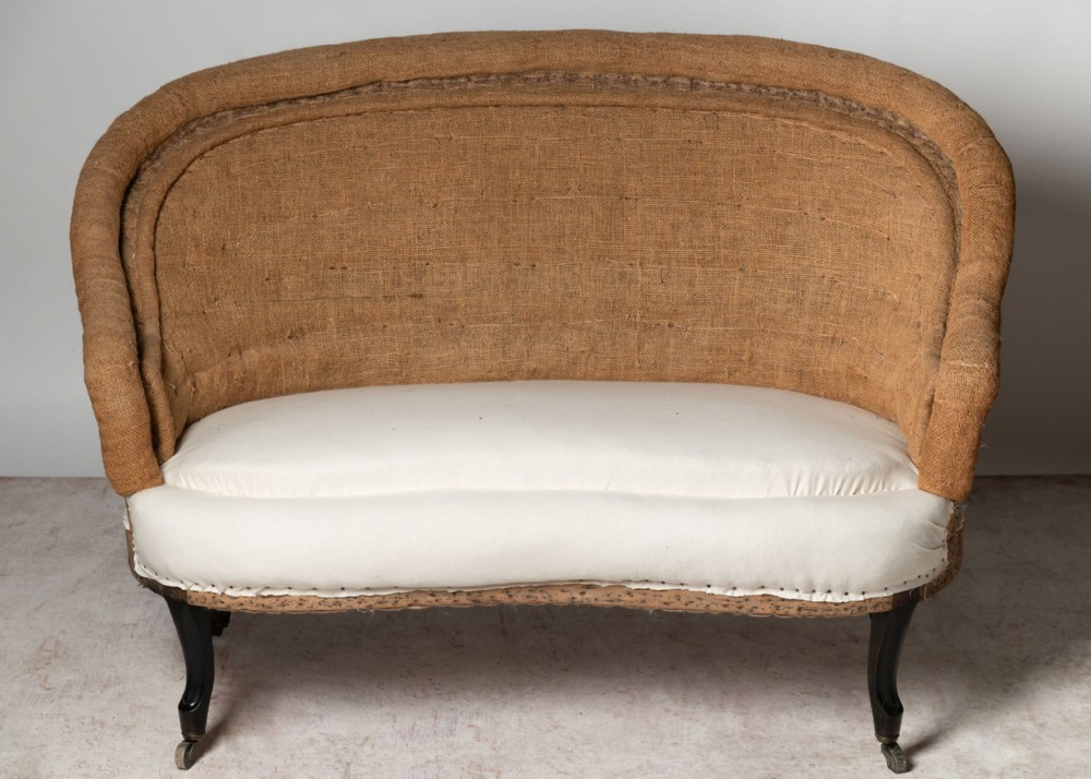 An antique French curved-back settee featuring exposed natural hessian upholstery, a white calico seat, and dark legs with brass castors.