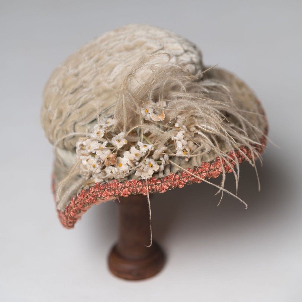 An antique Edwardian ladies hat made of ruched cream velvet, adorned with wispy feather plumes, small white cloth flowers, and a coral pink woven trim around the brim.