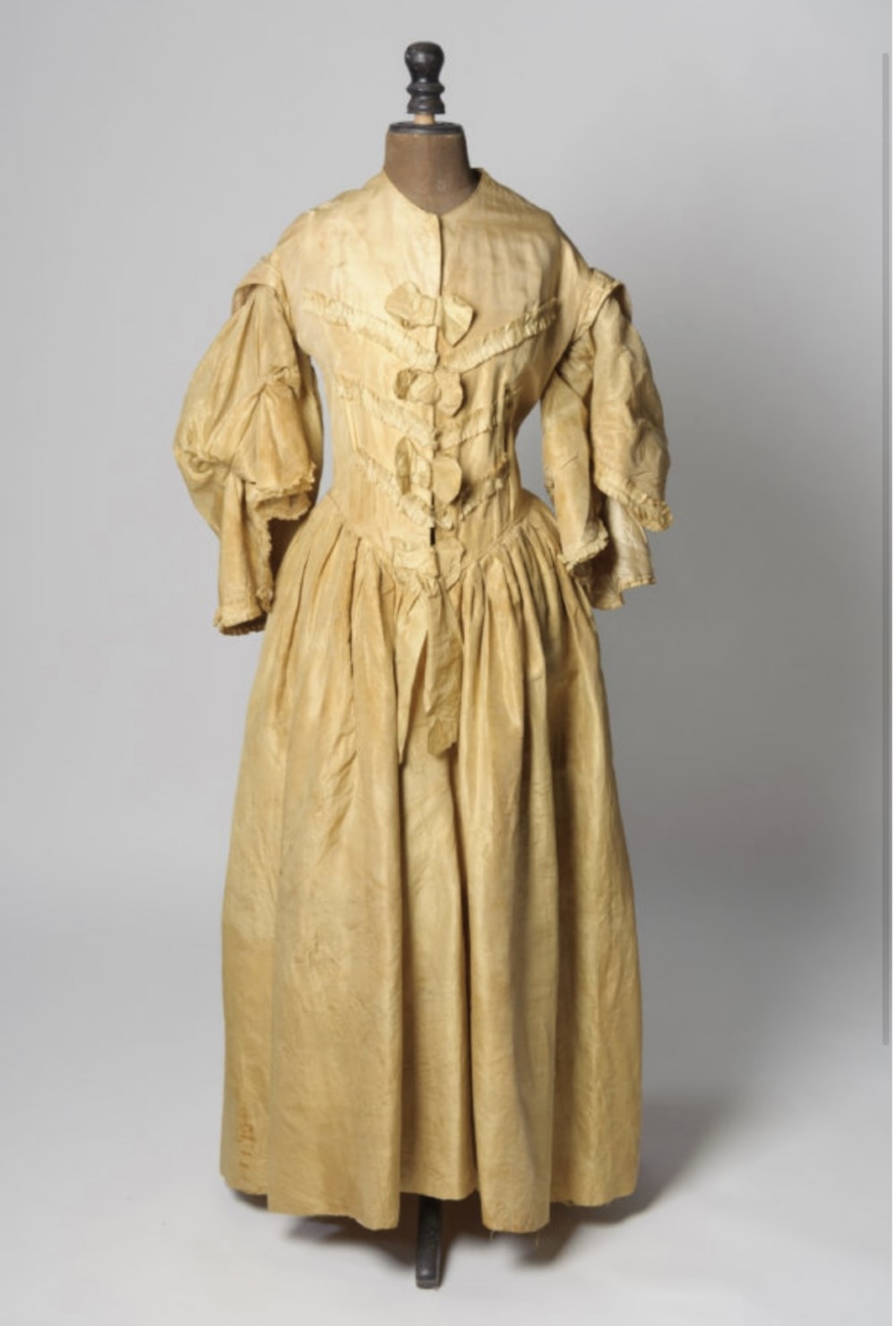 An antique champagne silk gown featuring voluminous gathered sleeves, a fitted boned bodice with front bow embellishments, and a full pleated skirt.