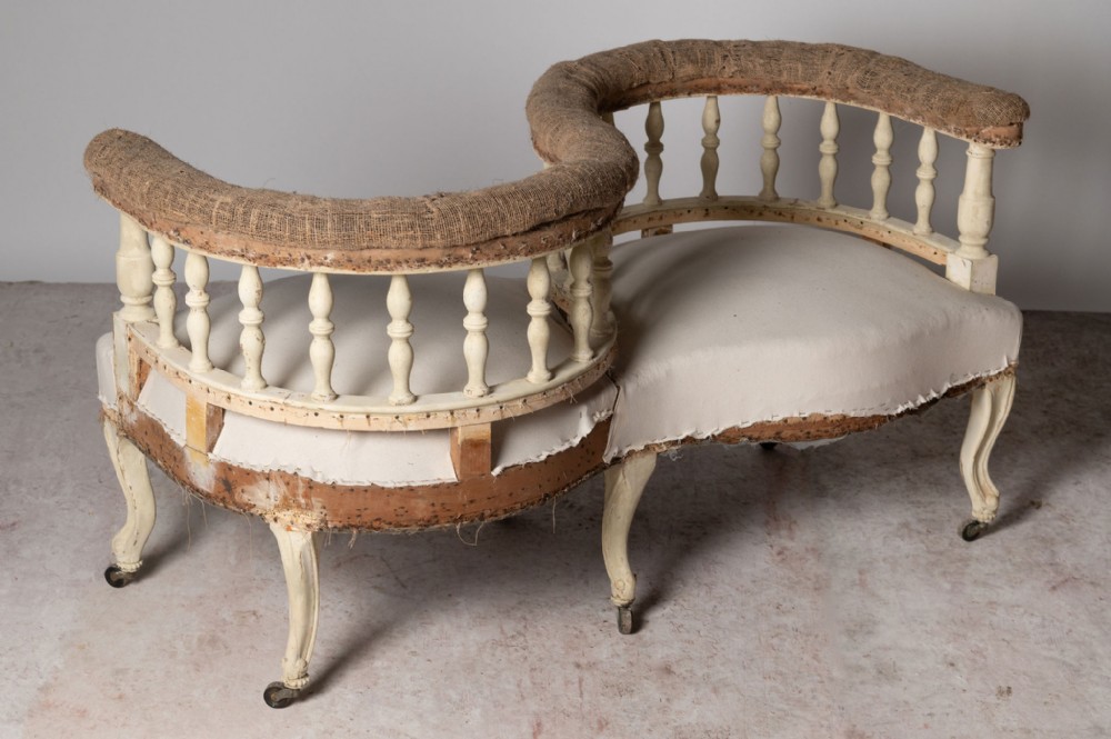 An S-shaped French tete-a-tete conversation sofa featuring a cream painted frame, turned spindles along the curved backrest, cabriole legs on castors, and exposed hessian upholstery.