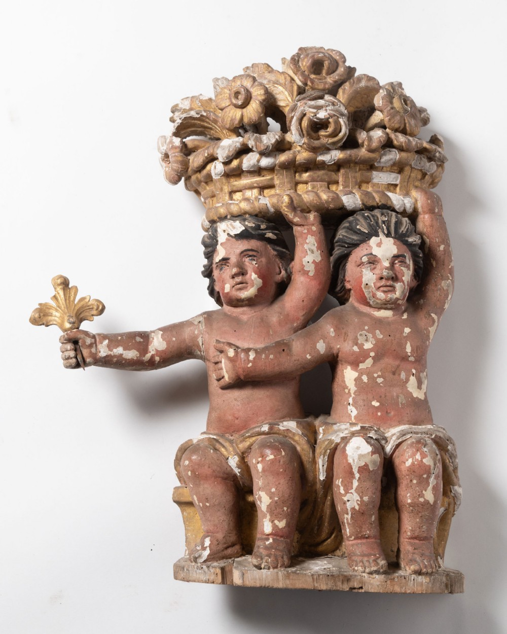 A carved wood polychrome and gilded relief sculpture featuring two seated putti supporting an elaborate basket of flowers on their heads.