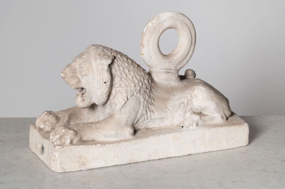 A recumbent plaster lion sculpture on an integrated rectangular plinth base featuring an open mouth and a circular loop handle on its back.