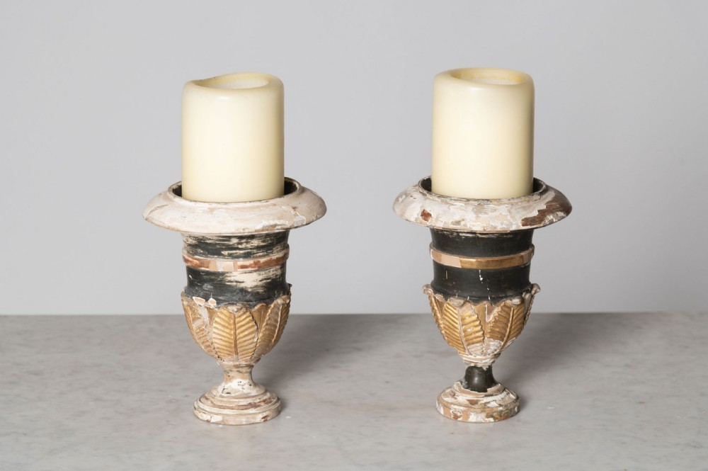 A pair of urn-shaped antique French wooden candlesticks featuring distressed black and off-white paint with gilded carved leaf decoration.