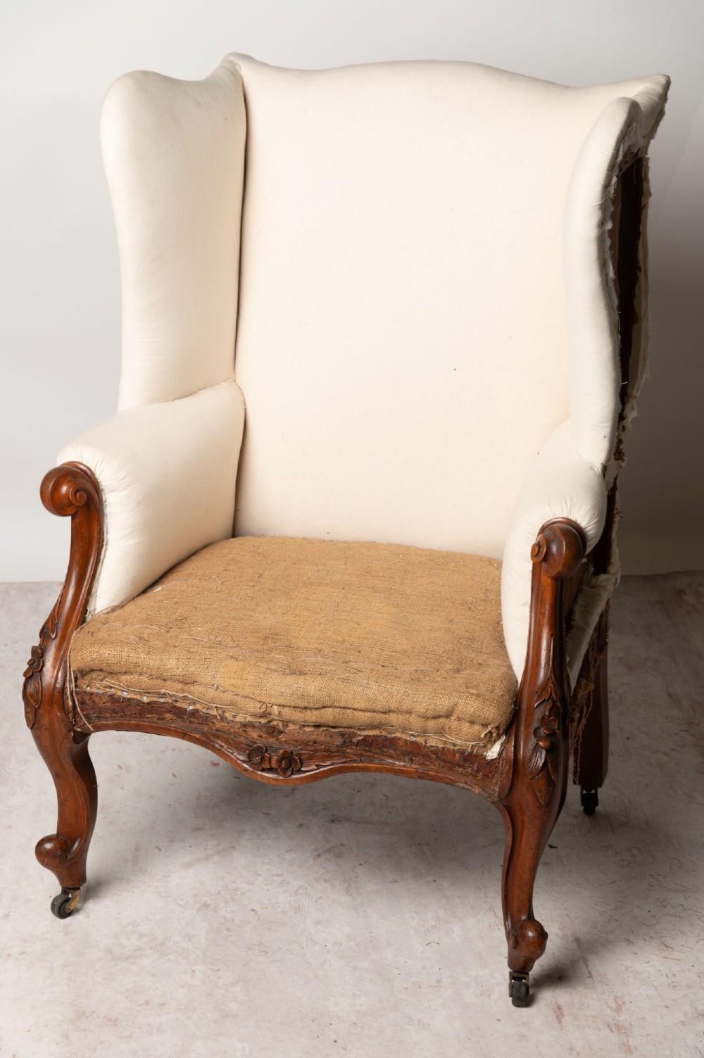 An antique English wingback armchair with carved wooden cabriole legs on castors, upholstered in cream calico with a hessian seat base ready for reupholstery.