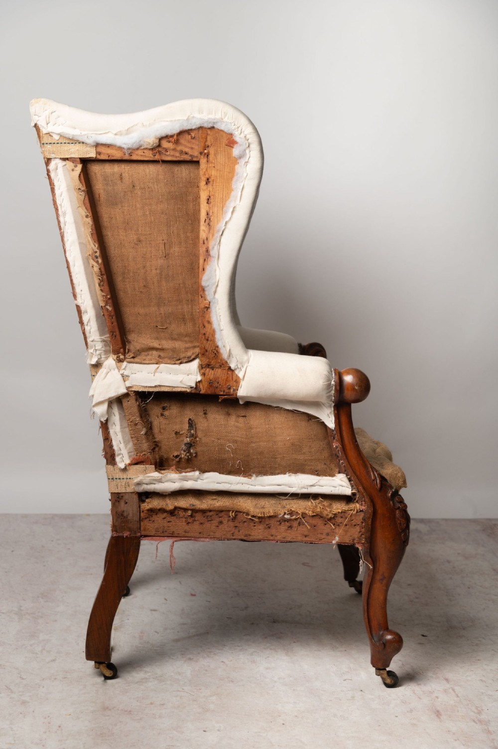 An antique English wingback armchair with carved wooden cabriole legs on castors, upholstered in cream calico with a hessian seat base ready for reupholstery. View II