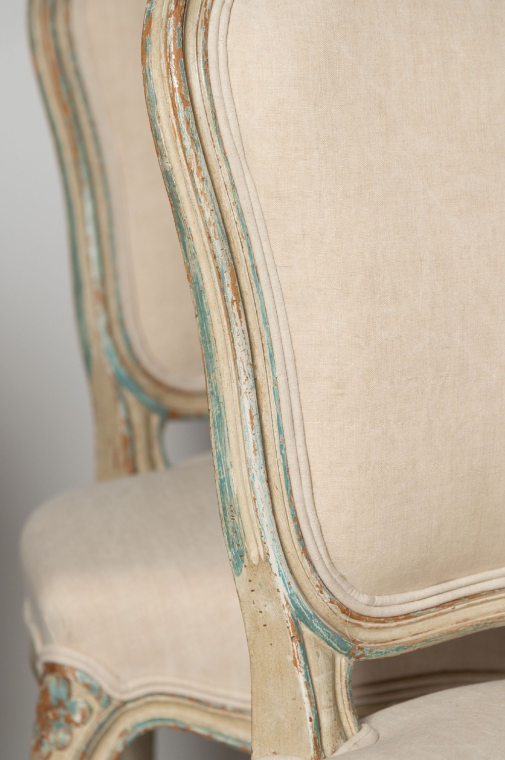 A pair of antique French side chairs featuring distressed off-white painted frames with floral crest carvings, cabriole legs, and neutral linen upholstery. View V