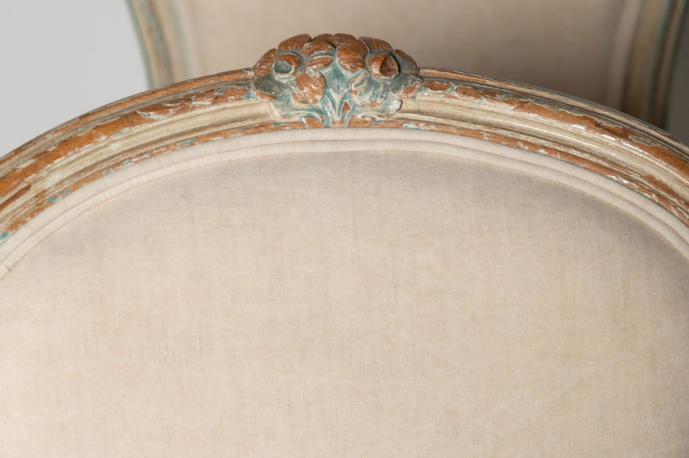 A pair of antique French side chairs featuring distressed off-white painted frames with floral crest carvings, cabriole legs, and neutral linen upholstery. View VI