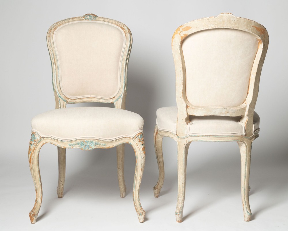 A pair of antique French side chairs featuring distressed off-white painted frames with floral crest carvings, cabriole legs, and neutral linen upholstery. View VIII